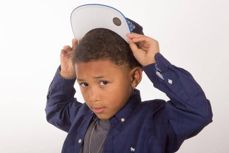 Young boy in blue shirt adjusting a snapback cap on his head against white background.