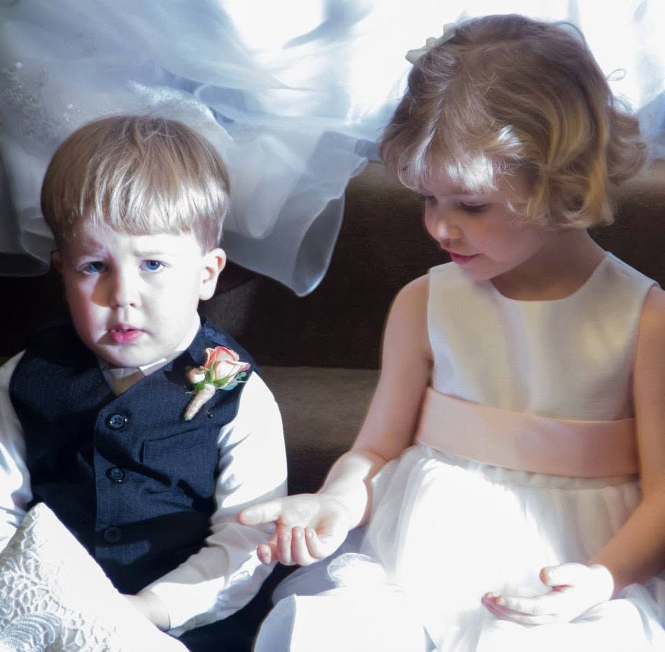 Young ring bearer in navy suit and flower girl in white dress sit together at a wedding ceremony.