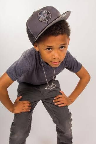 Young boy in gray cap, t-shirt, and pants poses confidently with hands on hips wearing a bat necklace.
