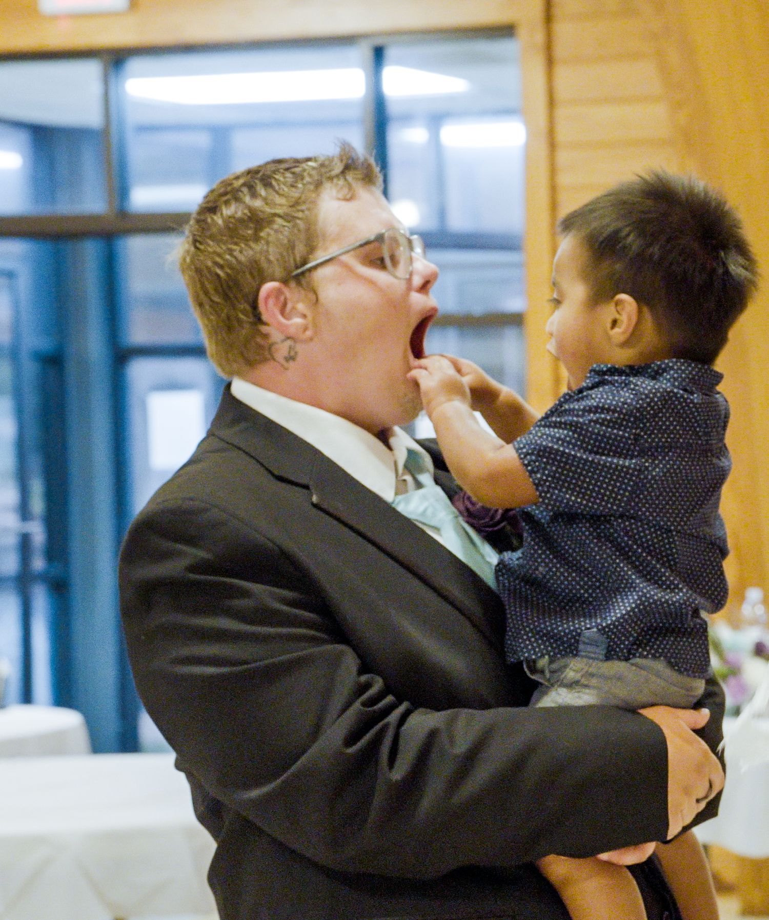 Adult in suit holding laughing toddler in blue dotted shirt, sharing a joyful playful moment indoors.