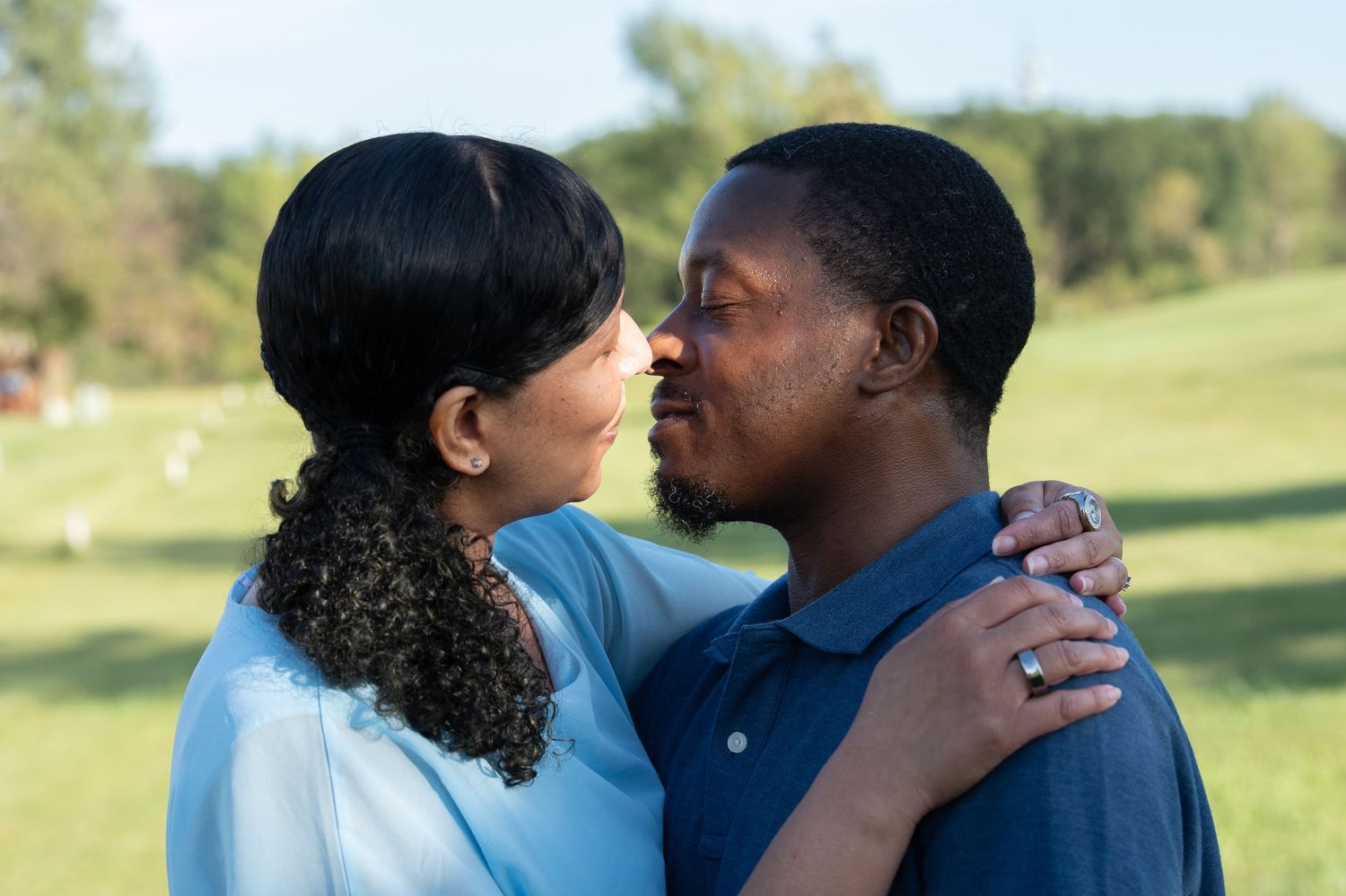 A couple shares a romantic moment outdoors, nearly kissing while embracing in a lush green park setting.