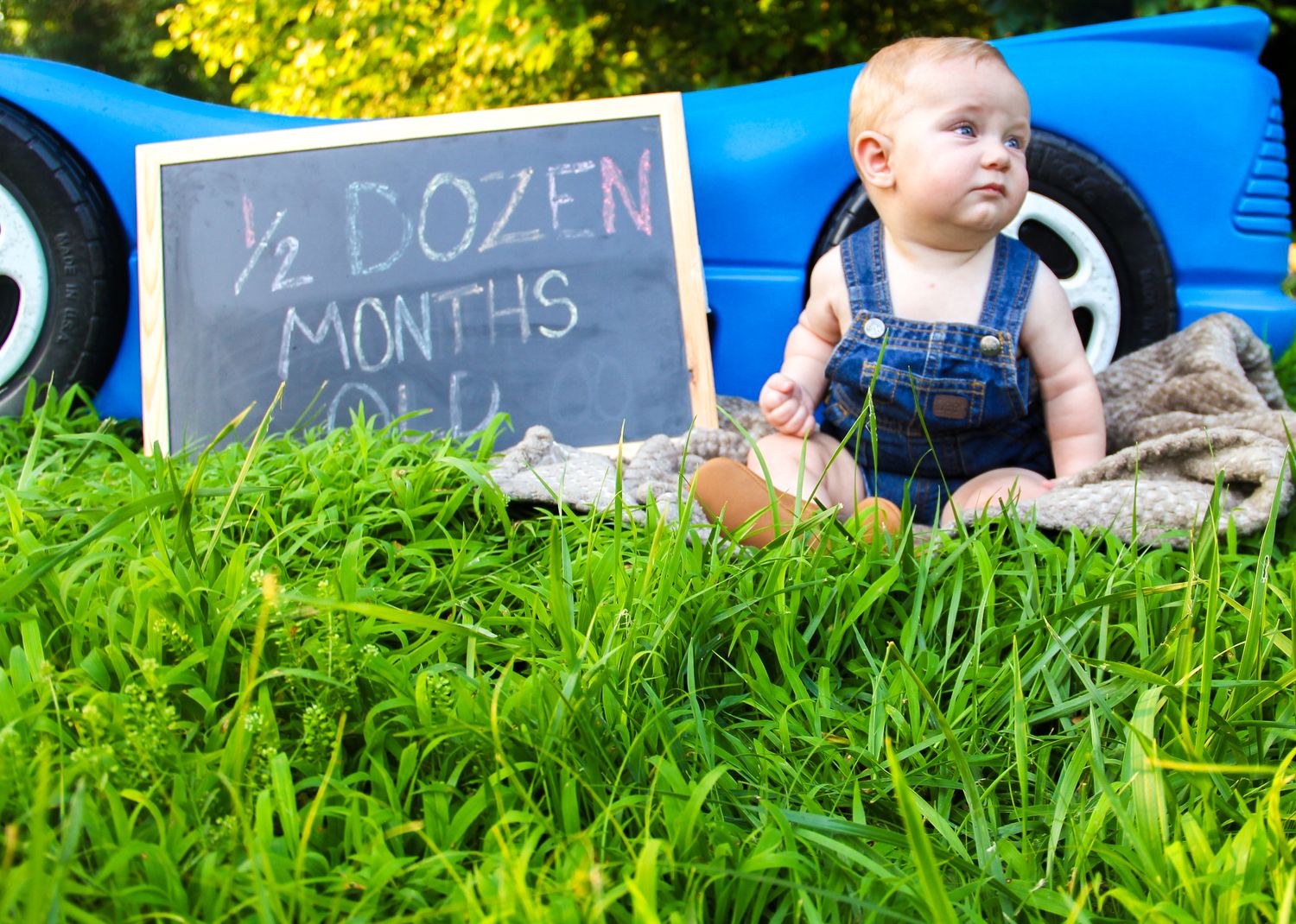 Baby in overalls sits in grass beside a chalkboard sign reading half dozen months old, leaning against a blue toy car.