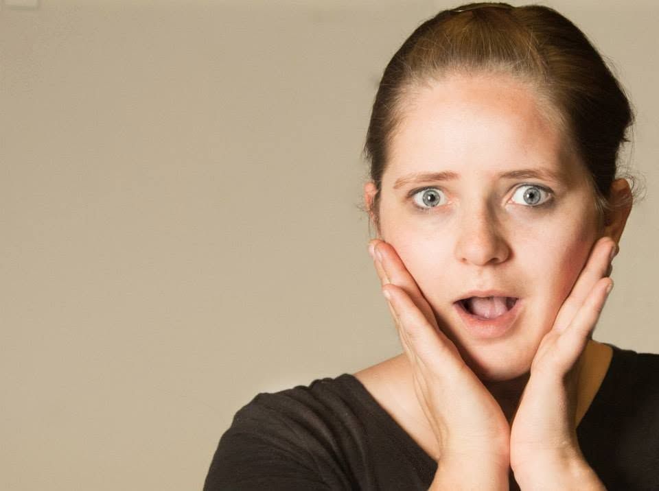 Surprised woman with blue eyes holds hands to cheeks with wide open mouth expression on grey background.