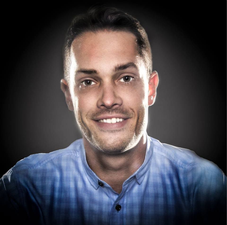 Smiling young man with short hair and stubble wearing a blue checkered shirt against a dark background.