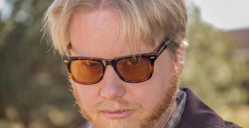 Blonde man wearing brown tortoiseshell sunglasses with orange tinted lenses outdoors in natural light.