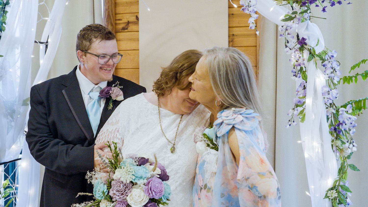 A joyful wedding ceremony with a groom in black suit and two women in white and floral dresses, surrounded by purple flowers.