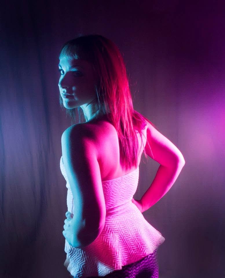 Young woman in pink peplum dress posed under dramatic neon blue and pink studio lighting.