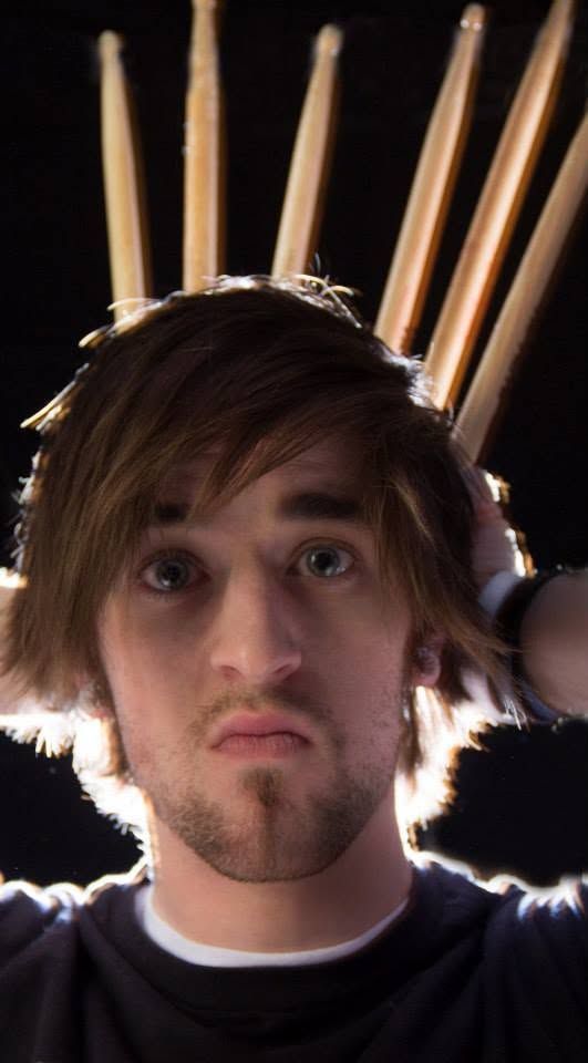 Creative portrait of a young man with drumsticks fanned behind his head against a dark background.