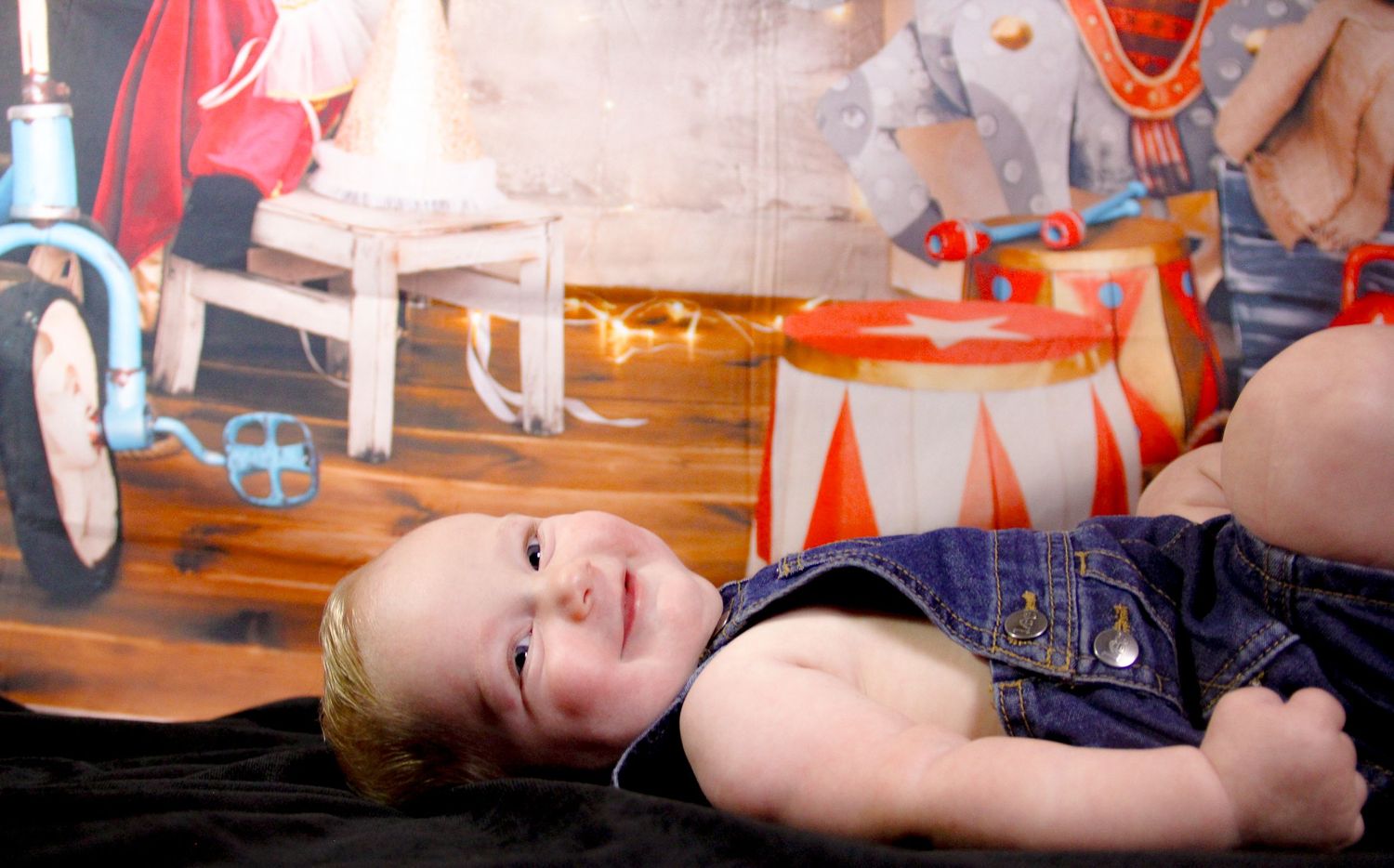 Smiling baby in denim overalls lying on floor in front of colorful circus-themed backdrop.