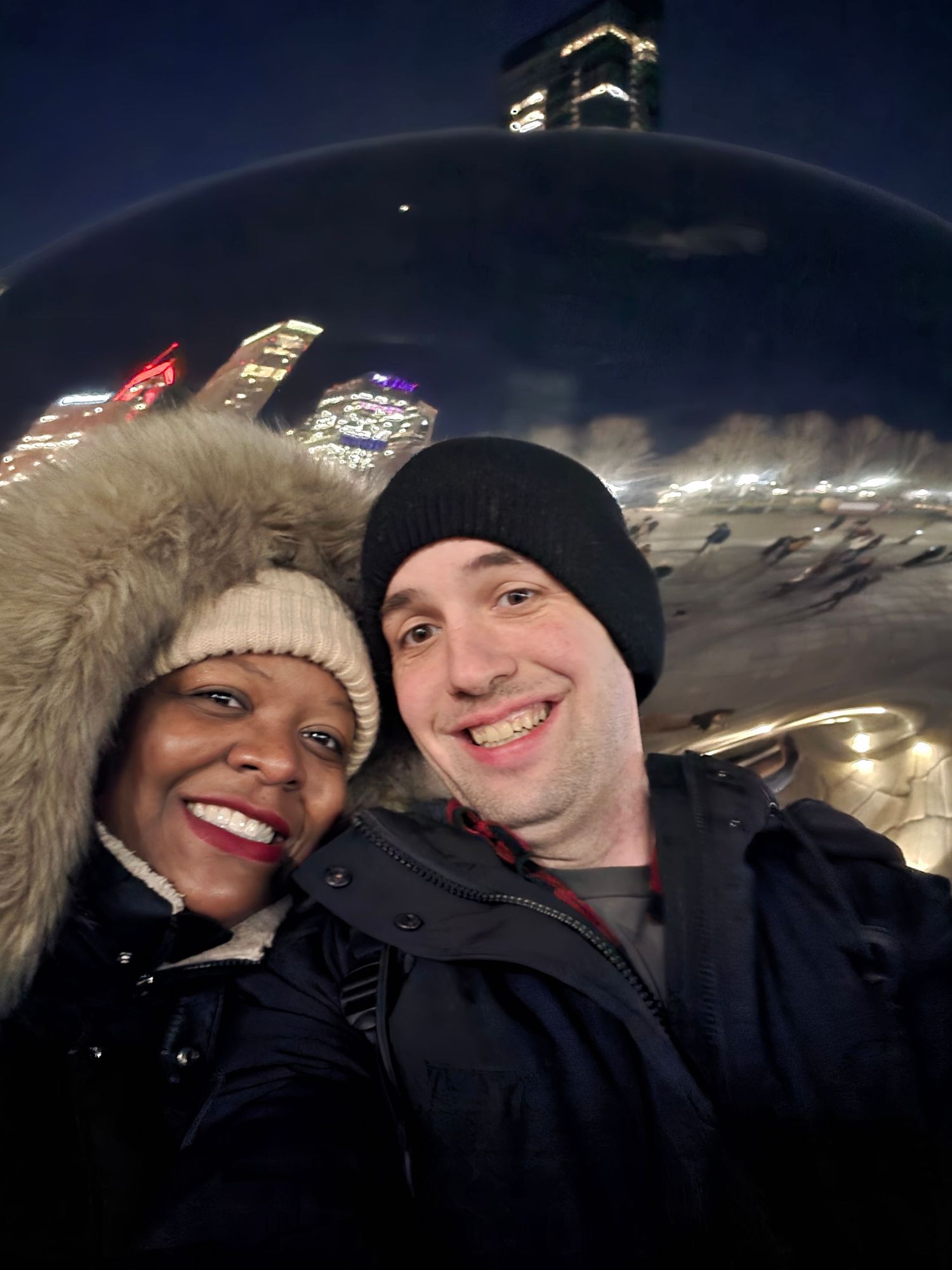 Couple smiling for selfie at night in front of Chicago Bean sculpture with city skyline reflected behind them.