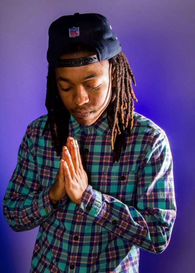 Young man with dreadlocks in plaid shirt and NFL cap poses with hands pressed together in prayer.