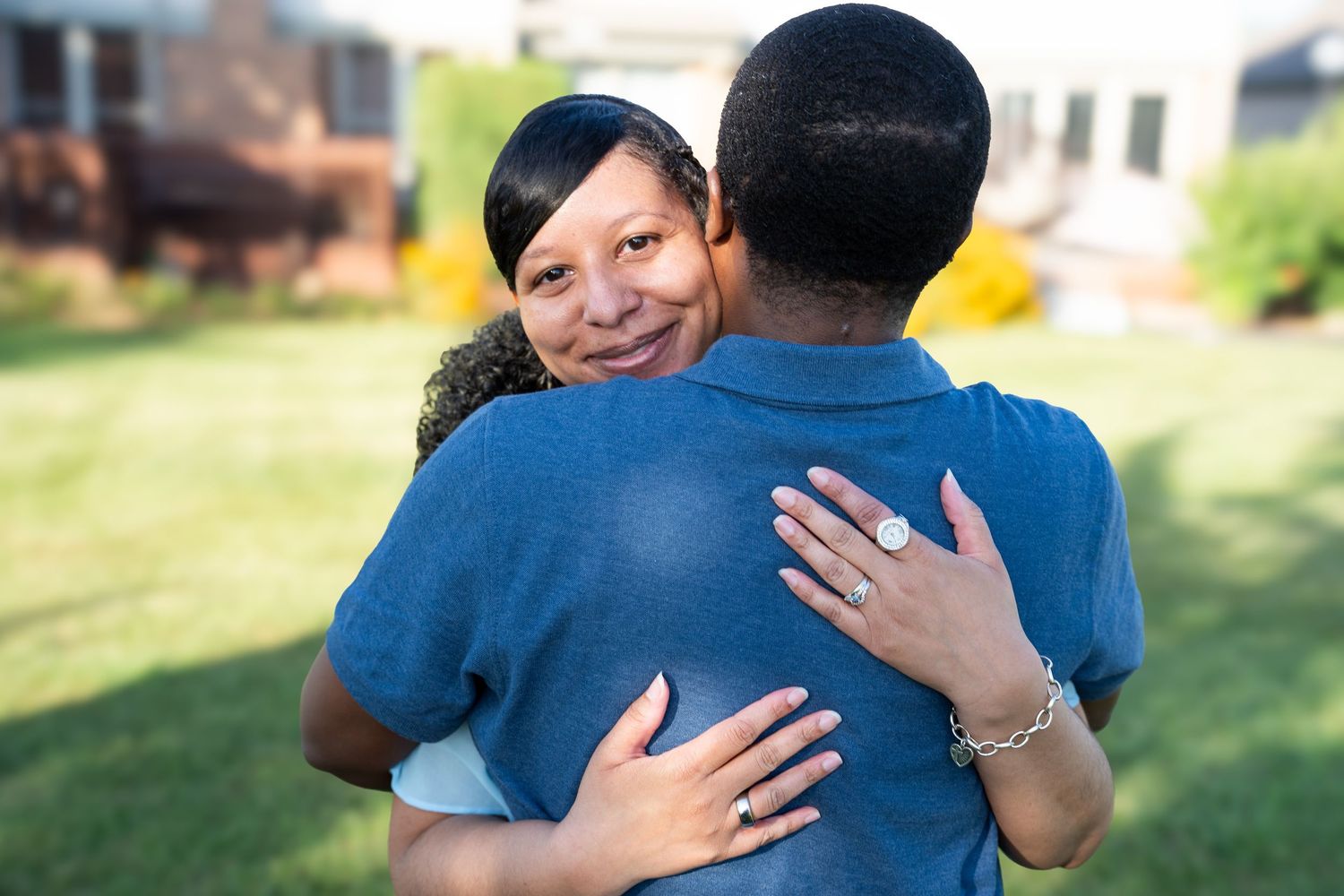 A woman with short hair smiles while hugging a person in a blue shirt outdoors in a green yard.