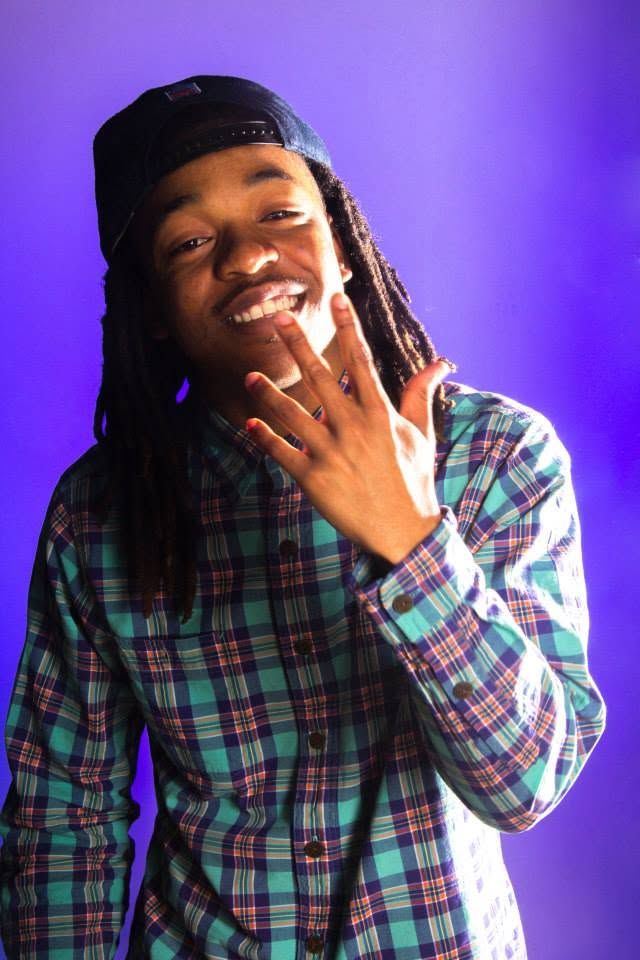 Smiling young man with dreadlocks wearing a backwards cap and plaid shirt against a purple background.