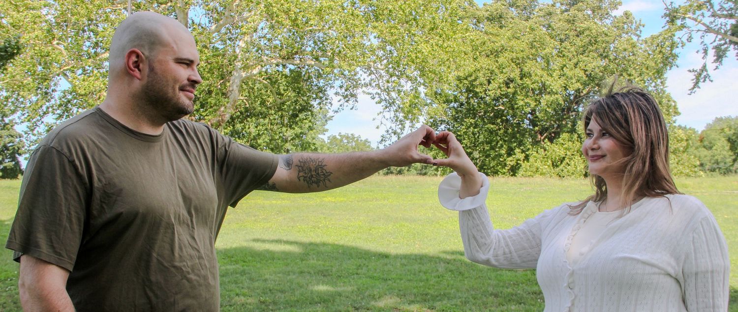 A couple holds hands outdoors in a green park, smiling at each other on a sunny day.