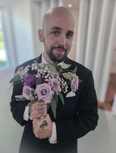 Bald man with beard in black suit holding purple rose bouquet at wedding ceremony.