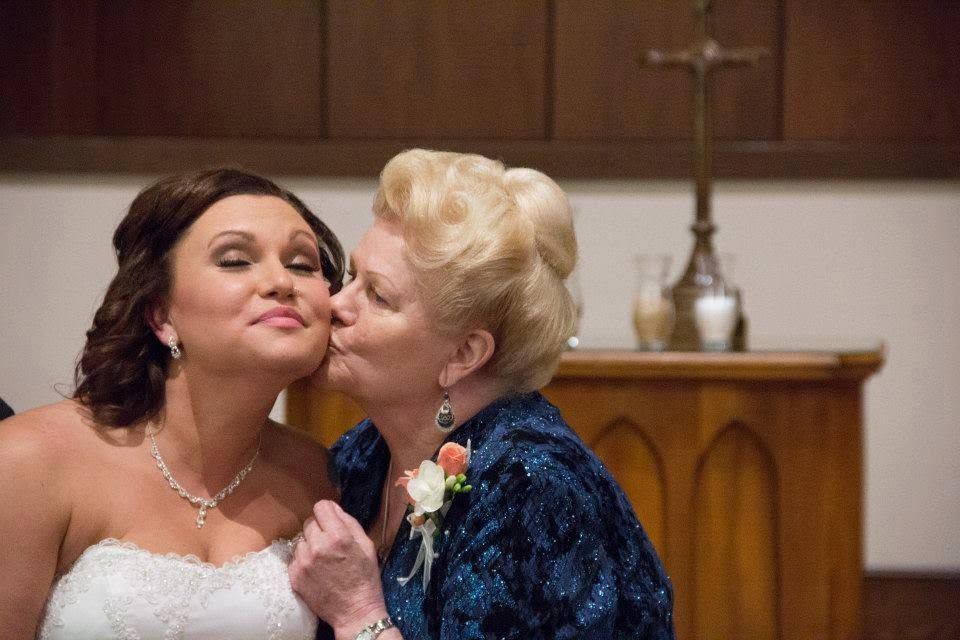 Bride in white gown receives a loving kiss on the cheek from an older woman in a blue dress inside a church.