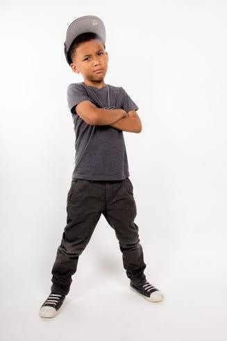 Cool young boy in gray t-shirt, dark pants, and snapback cap posing with arms crossed in sneakers.