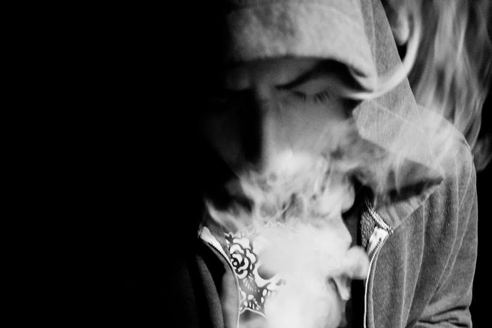 Black and white photo of a hooded person exhaling smoke, with a decorative pendant visible.