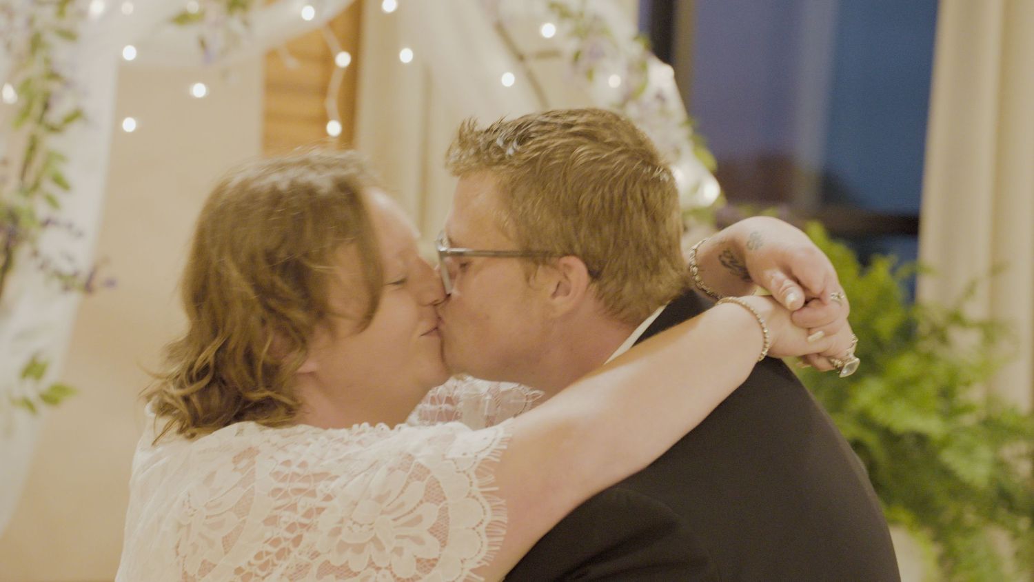 A couple shares a romantic first kiss at their wedding ceremony, with the bride in lace and groom in a suit.