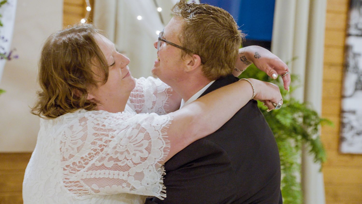 Two women share a warm embrace and smile at each other during a wedding ceremony indoors.