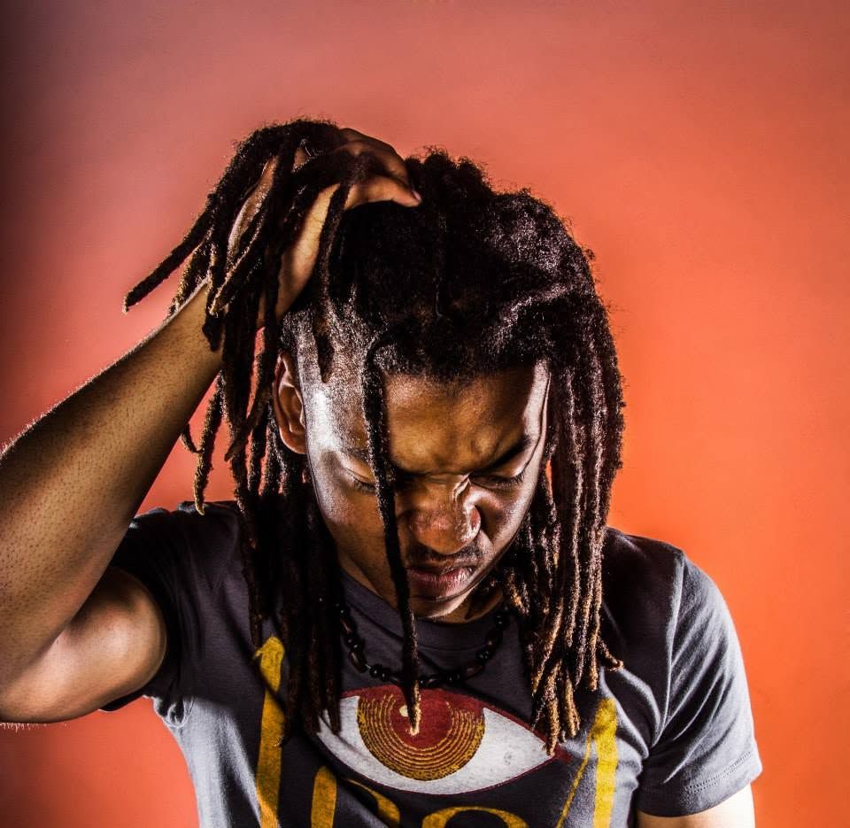 Man with long dreadlocks wearing graphic tee posed against vibrant orange background.