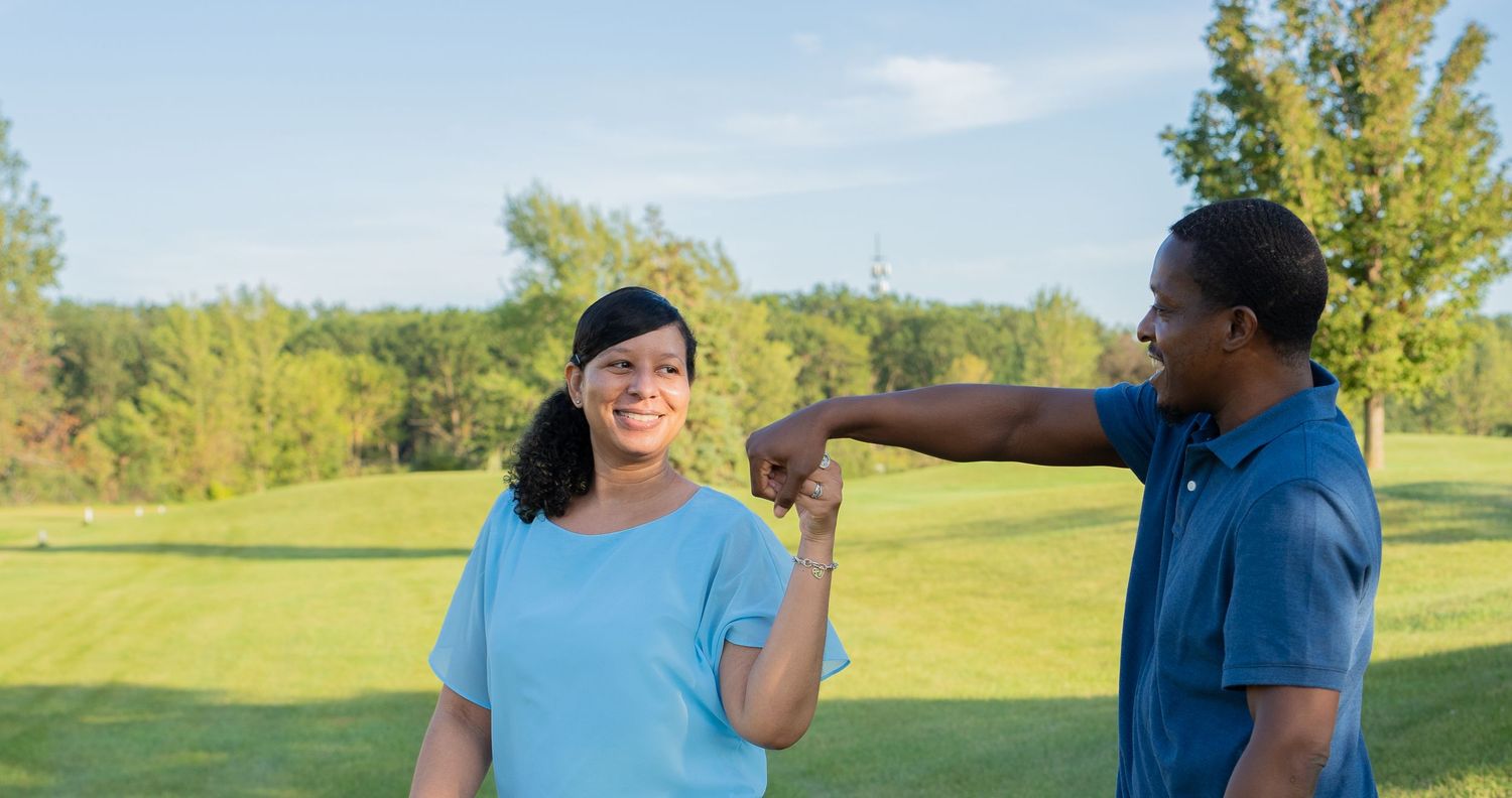 A woman in a light blue shirt fist bumps a man in a blue polo on a sunny green golf course.