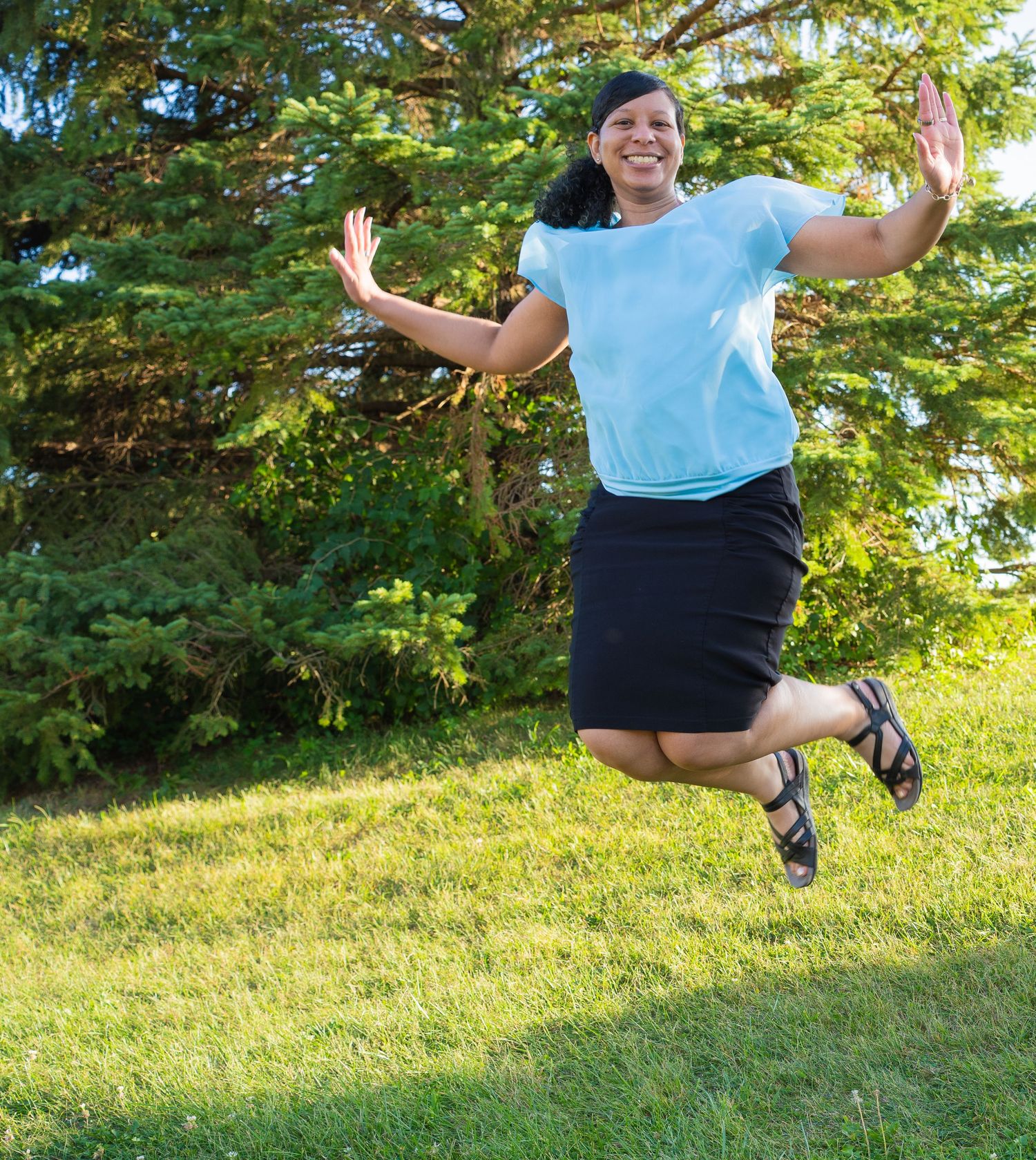 A joyful woman in a light blue shirt and black skirt jumps energetically on green grass with trees behind her.