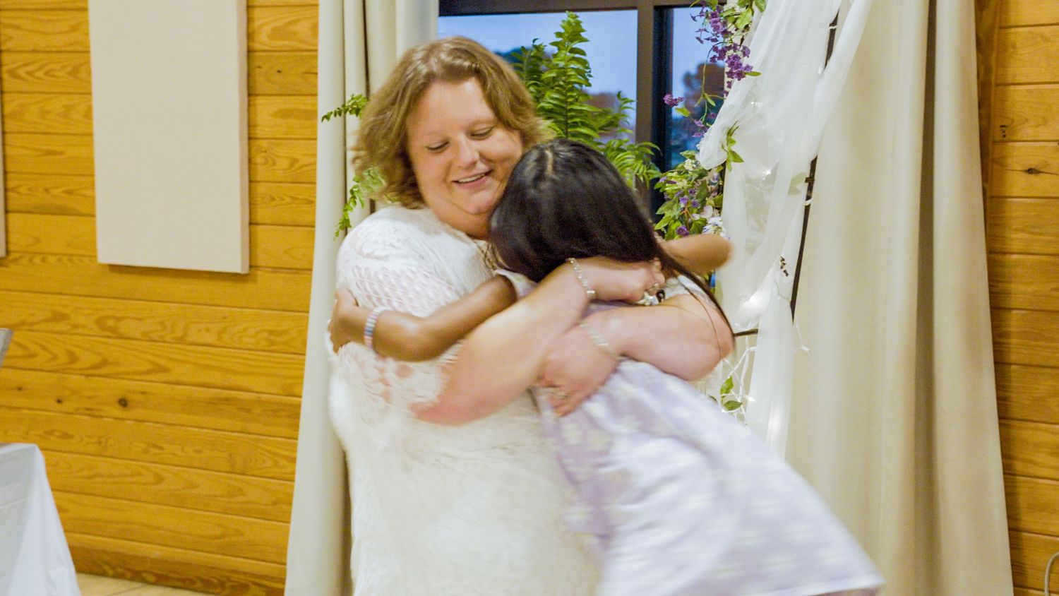 Two women in white dresses share a warm hug at a wedding venue with wood-paneled walls.
