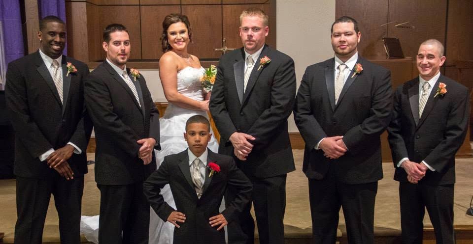 Wedding party poses formally with bride in white gown, groom, groomsmen in black suits, and young boy in church.
