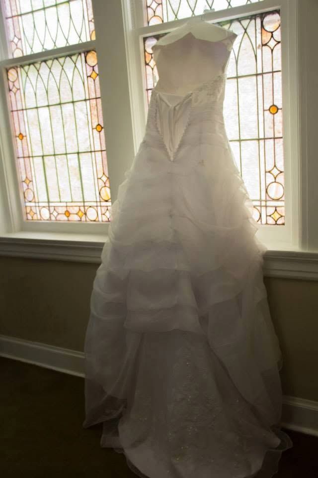 White strapless tiered wedding gown hanging in front of stained glass church windows with natural light.