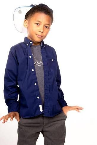 Young boy in navy blue button-down shirt and grey jeans poses confidently with a snapback hat tilted back.