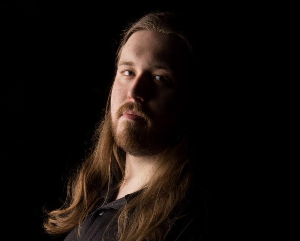 Man with long auburn hair and beard against dark background, dramatic studio portrait photography.