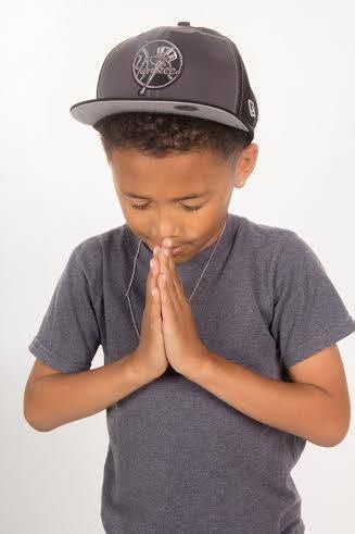 Young boy in gray t-shirt and cap with hands pressed together in prayer pose against white background.