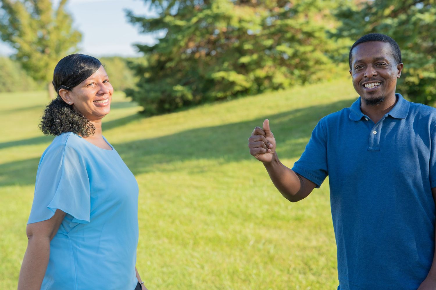 Two people smiling outdoors in a park, man giving thumbs up wearing blue polo, woman in light blue shirt on green lawn.