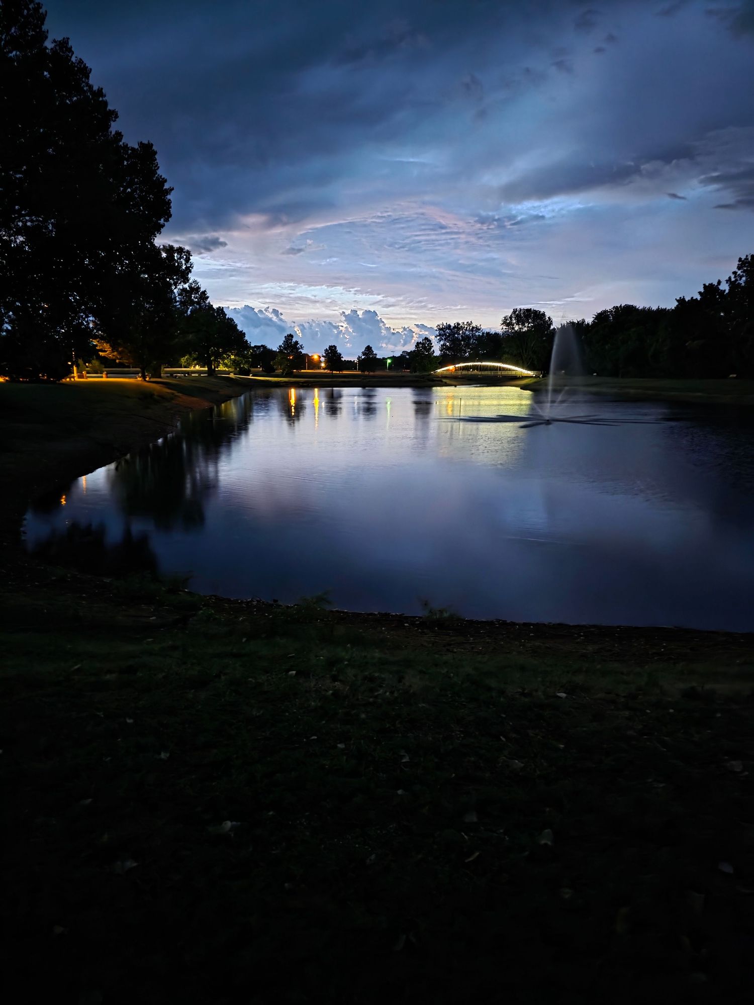 Twilight view of a serene park pond with a illuminated arched bridge, fountain, and dramatic cloudy sky reflected on water.