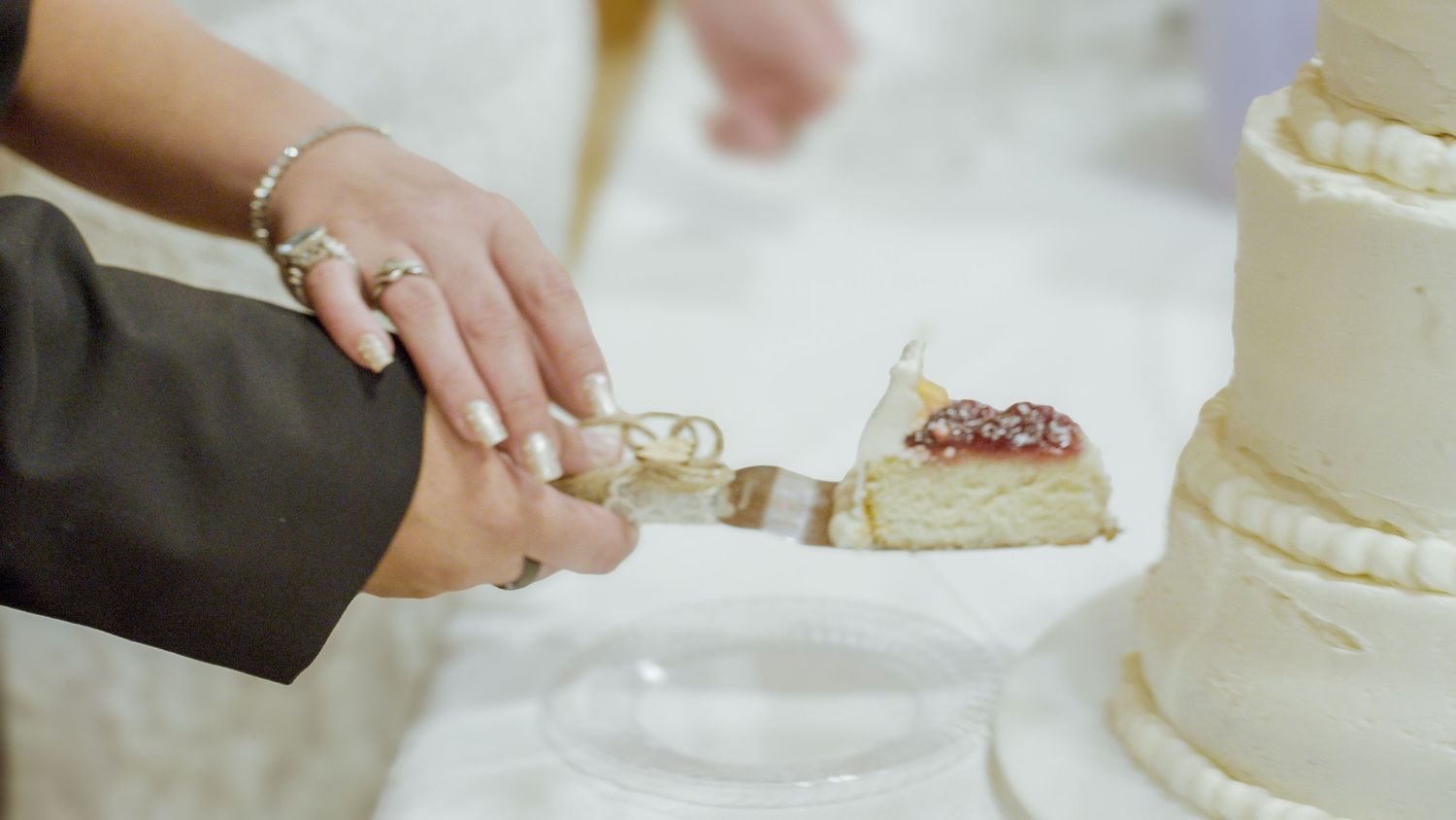 Bride and groom cutting a white tiered wedding cake together, with elegant decorations and rings visible.