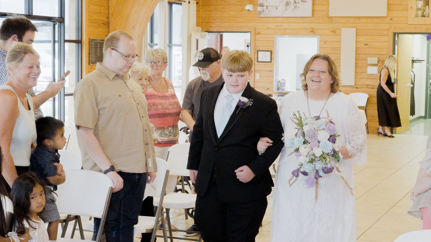 A bride in a white dress walks down the aisle with a young man in a tuxedo during an indoor wedding ceremony.