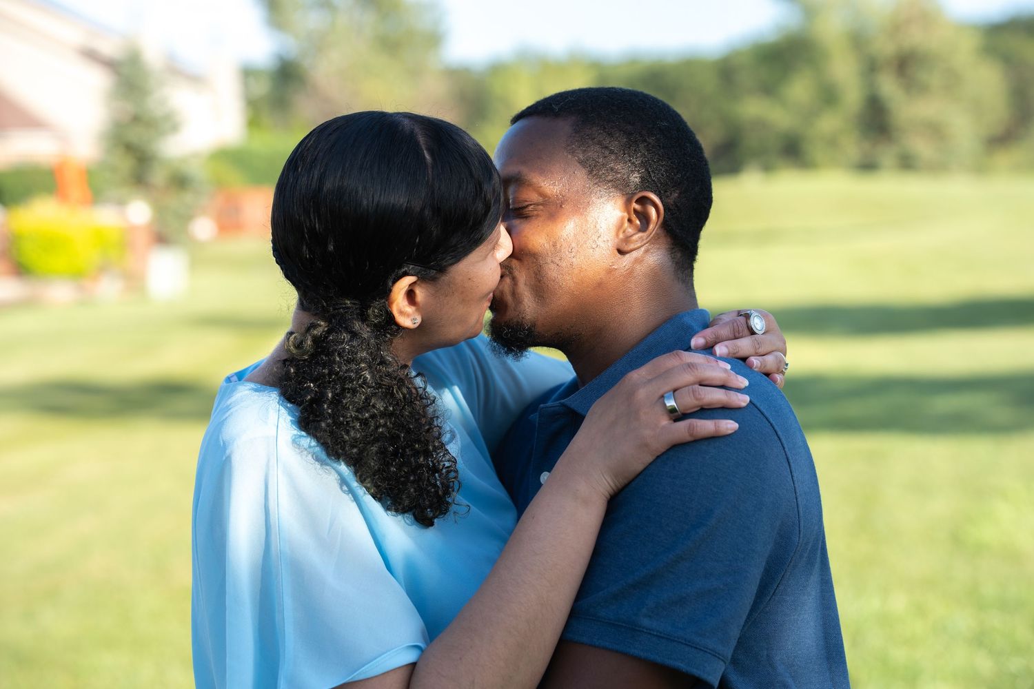 A couple sharing a romantic kiss outdoors on a green lawn near a house.