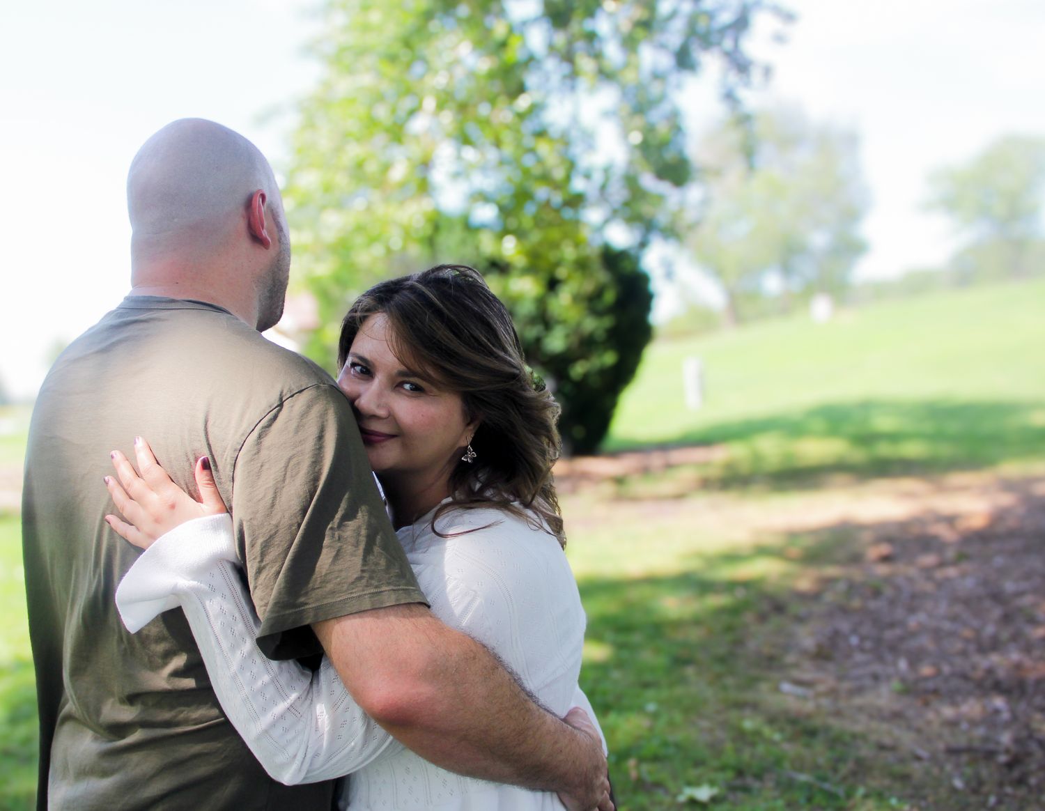 Couple embracing outdoors in a park, woman smiling over man's shoulder in a romantic portrait.