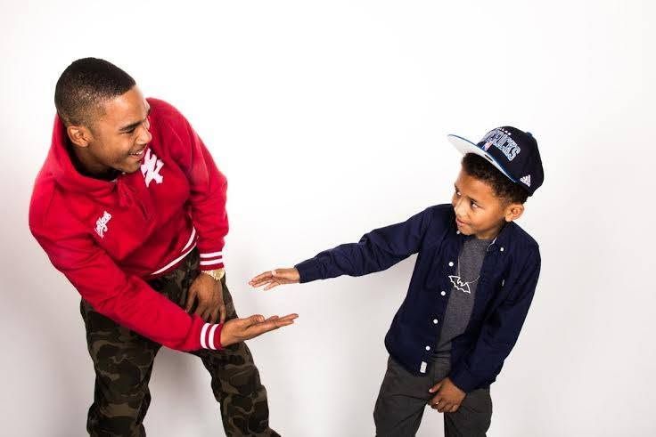 A man in a red jacket and young boy in a navy cap share a playful handshake gesture against white background.