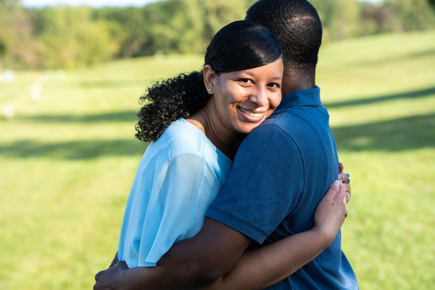 A smiling Black woman hugs a man outdoors in a green park, wearing a light blue top.
