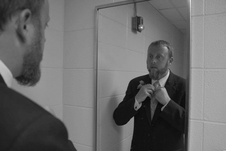 Black and white photo of a bearded man in a suit adjusting his tie in a bathroom mirror.