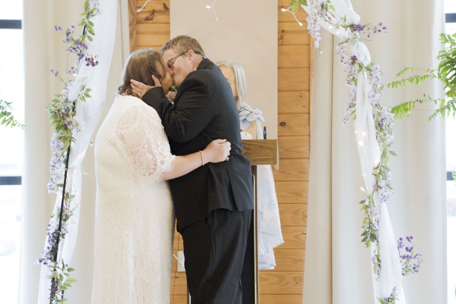 Bride and groom share first kiss at wedding altar decorated with white fabric and purple floral garlands.