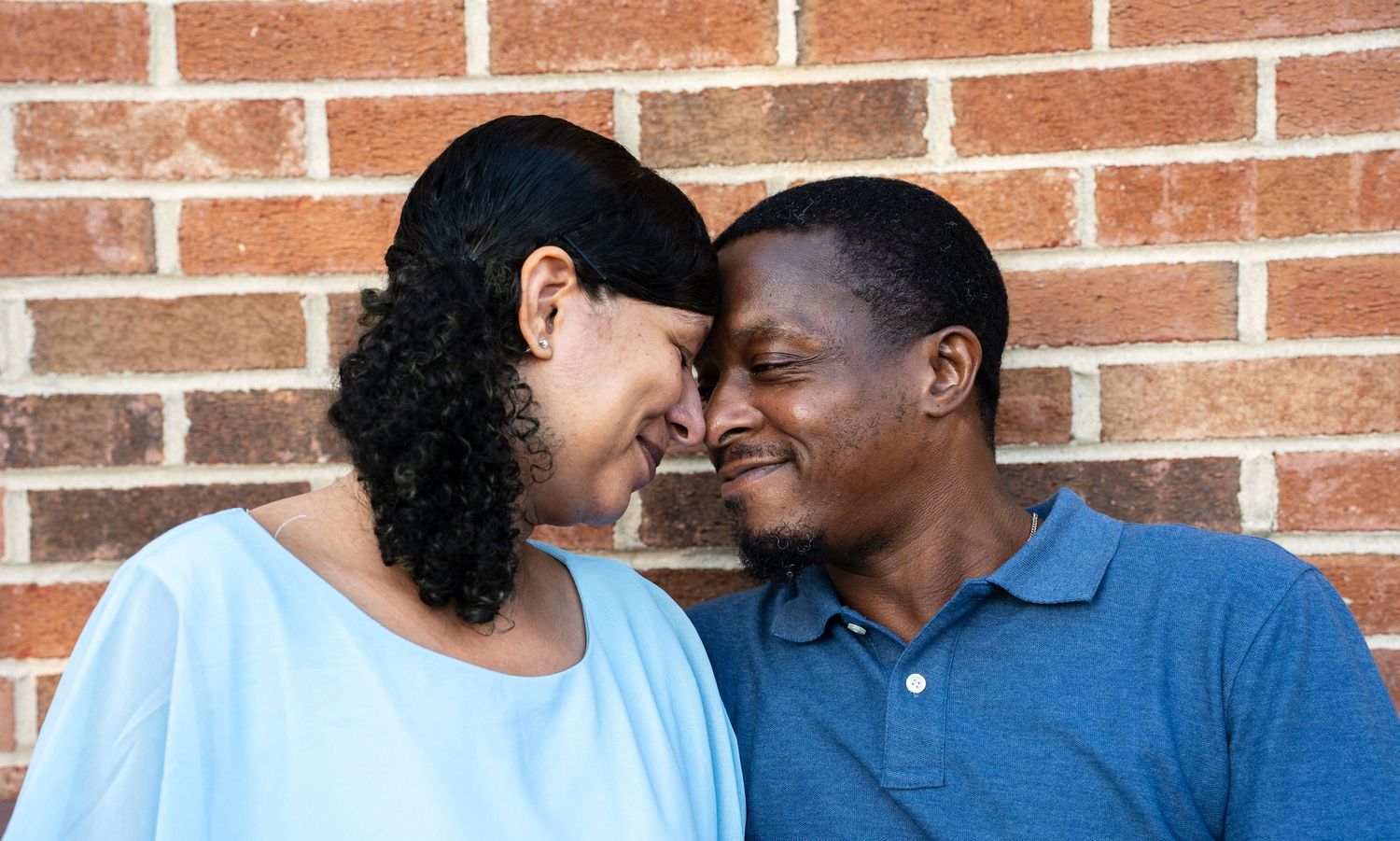 A loving Black couple shares an intimate moment, touching foreheads and smiling against a brick wall.