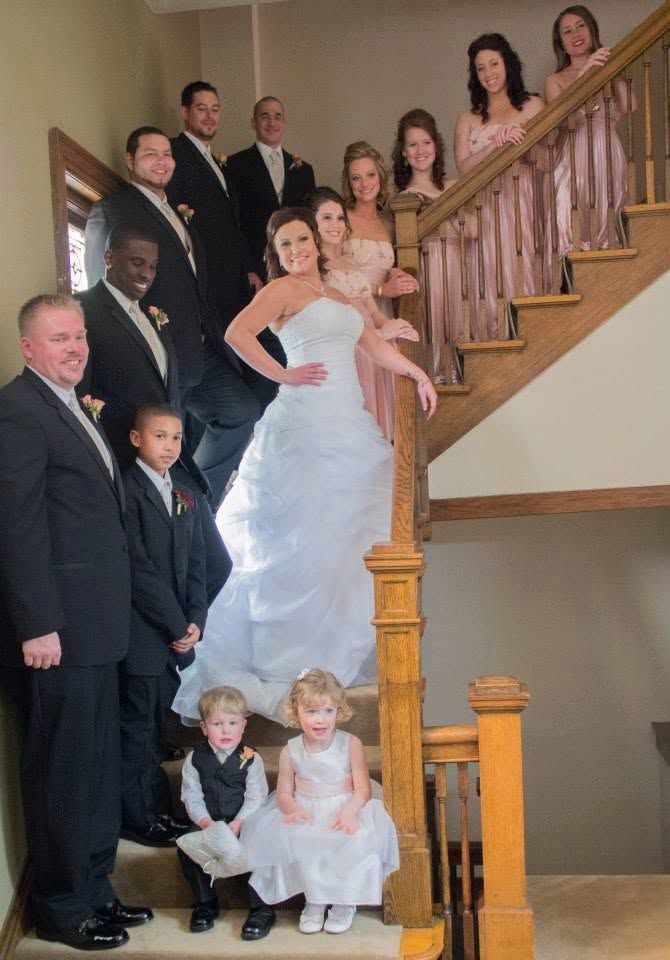 Wedding party poses on staircase with bride in white gown, bridesmaids in pink, groomsmen in suits, and two young children.