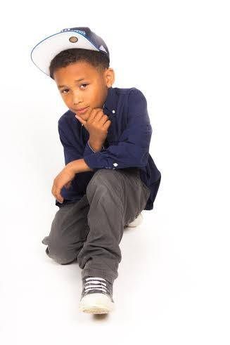 A young boy in a navy shirt, gray pants, and snapback cap sits thoughtfully on a white studio background.