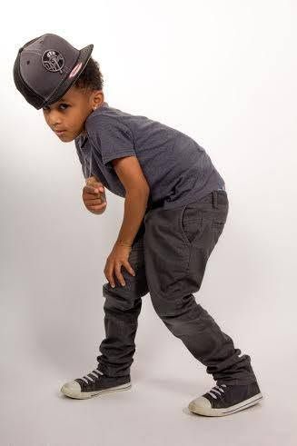 Young boy in hip-hop style outfit with gray joggers, sneakers, and snapback cap striking a cool dance pose.