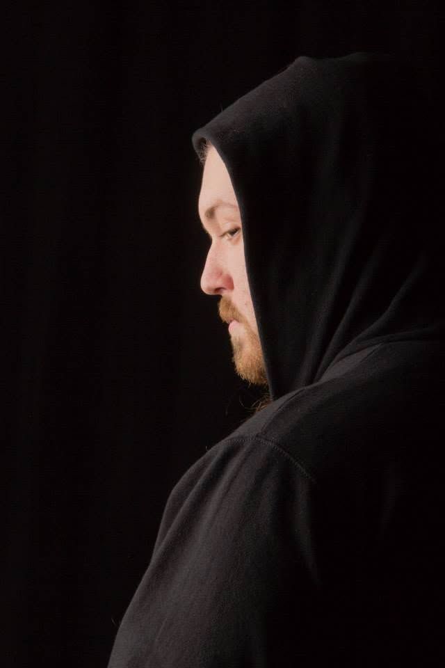 Side profile of bearded man wearing black hooded sweatshirt against dark background.