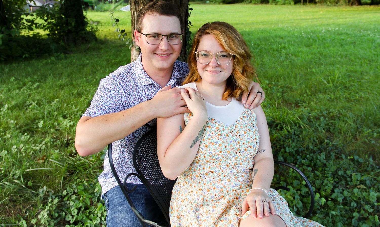 Young couple smiling outdoors, sitting together on grass near a tree in a sunny green park setting.