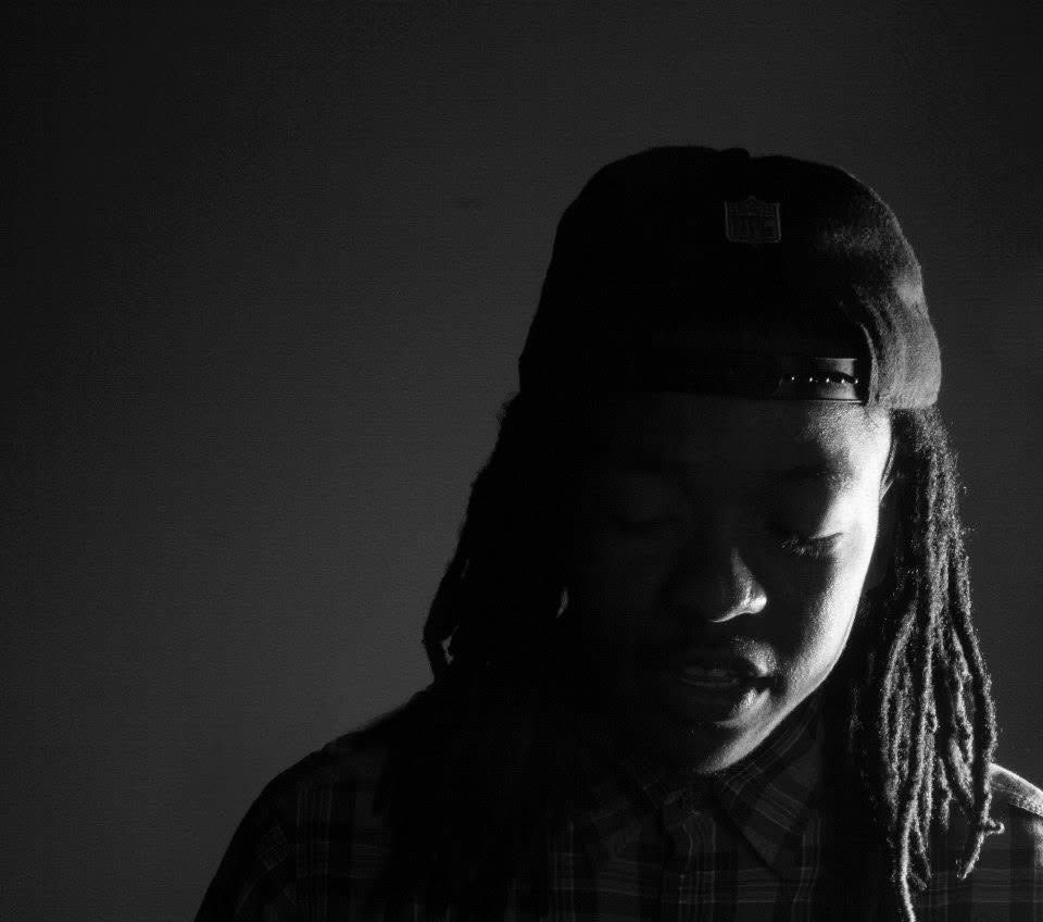 Black and white portrait of a young man with dreadlocks wearing a backwards cap, looking downward.